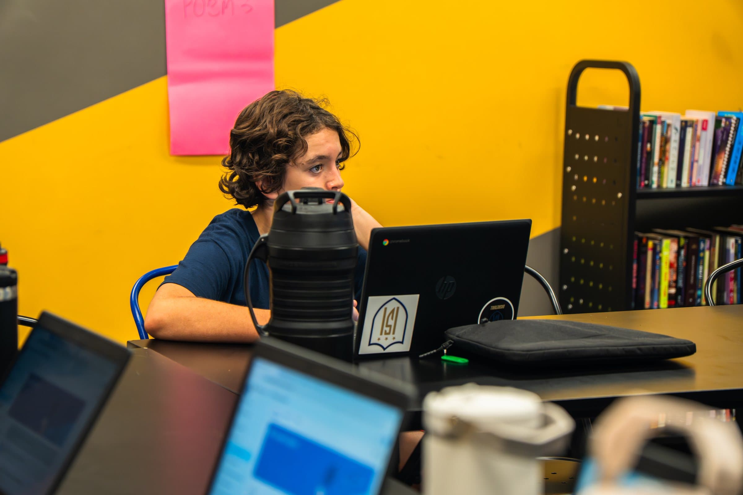 A student in an ISI Academy shirt working at a laptop in a bright classroom.